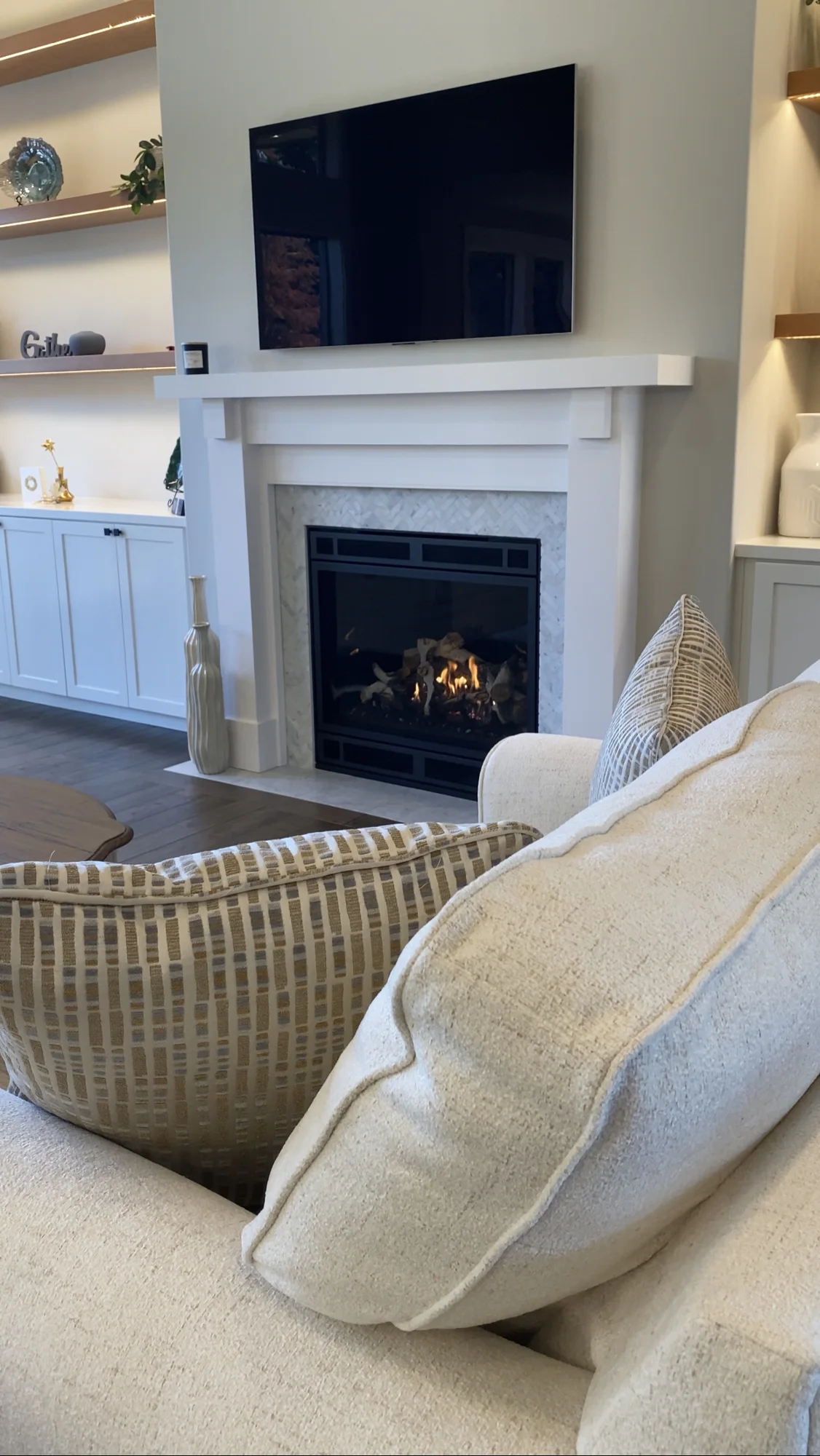 Living room with a cozy fireplace, beige couch with patterned cushions, and a TV mounted above the mantel. Shelving with decorative items flanks the fireplace.